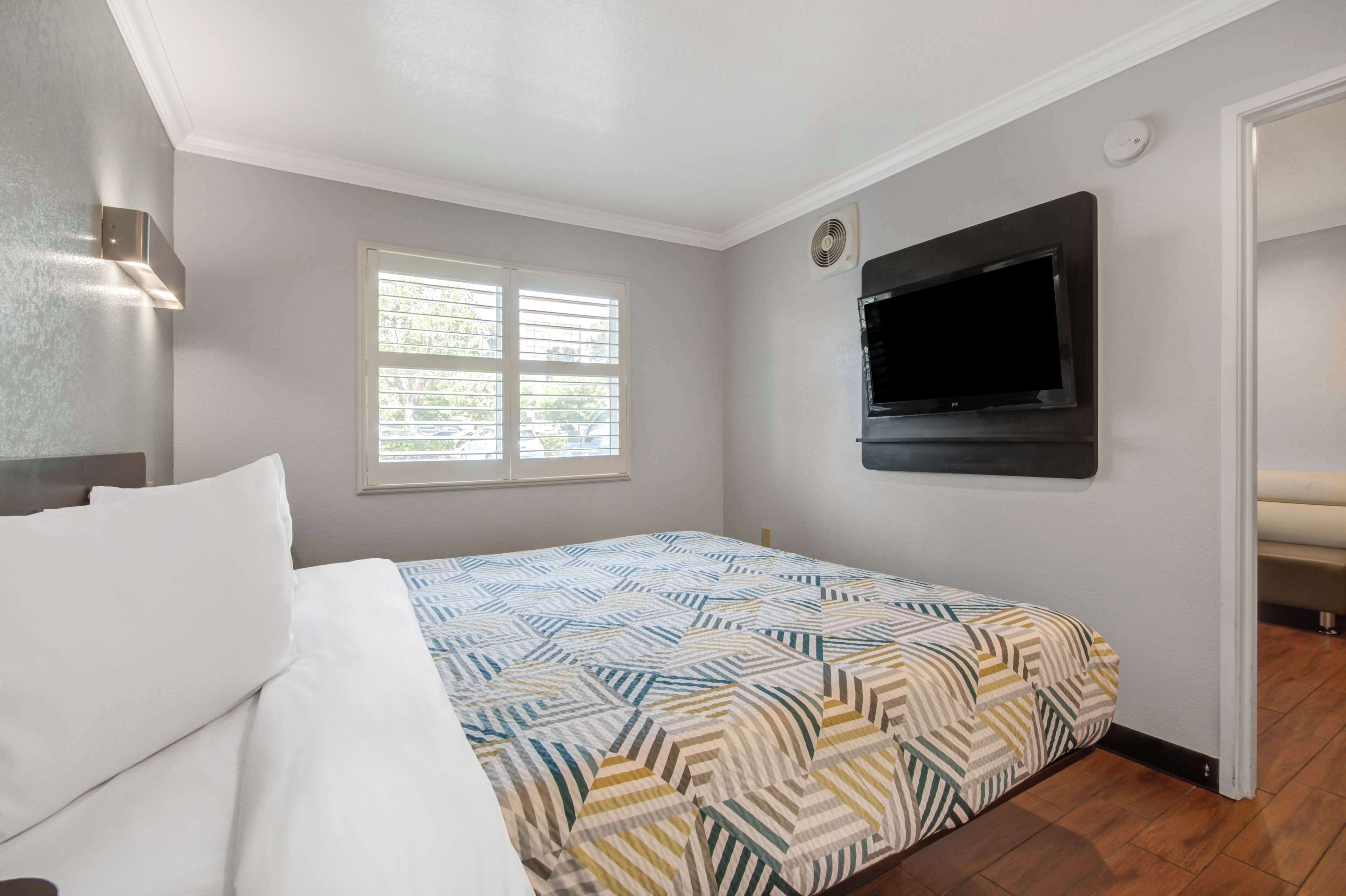 A bedroom with a bed, a window with blinds, a flat-screen TV mounted on the wall, and a ceiling fan.