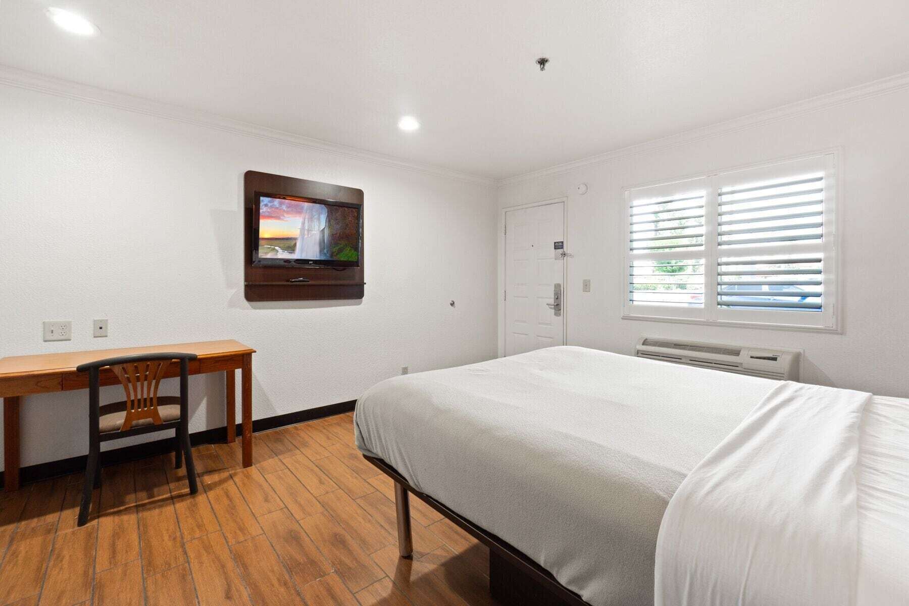A hotel room with a bed, a desk, a chair, a TV mounted on the wall, a door, and a window with blinds.