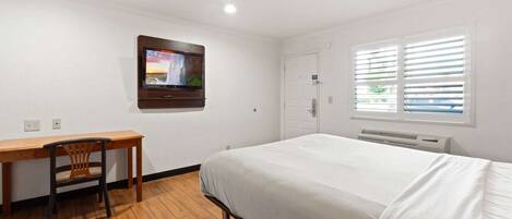 A hotel room with a bed, a desk, a chair, a TV mounted on the wall, a door, and a window with blinds.