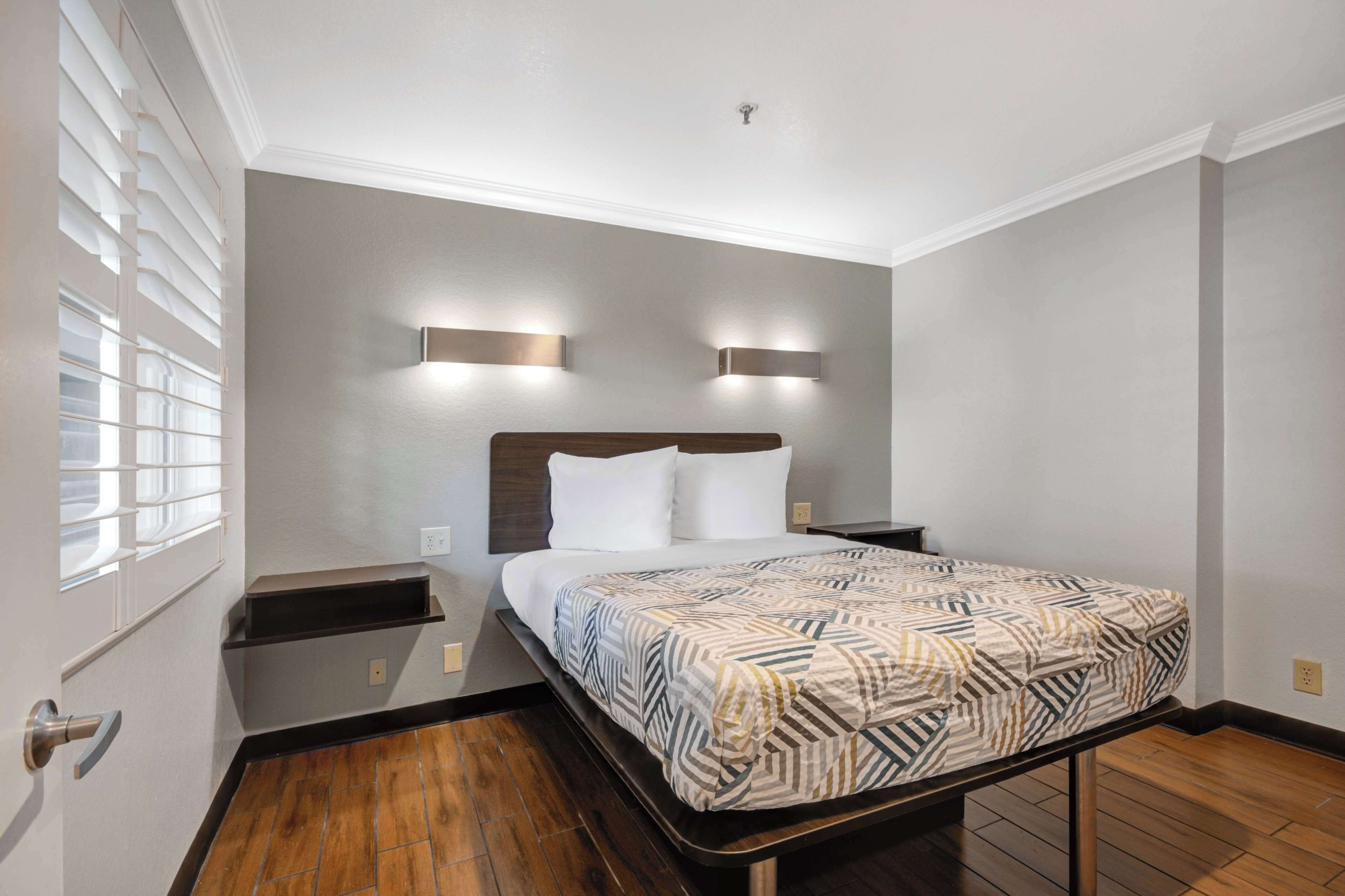 A hotel room with a bed, bedside tables, a window with blinds, and wall-mounted lighting.