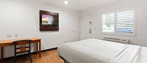 A hotel room with a bed, a desk, a chair, a television mounted on the wall, and a window with blinds.