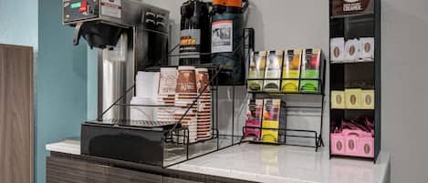 A coffee station with a coffee machine, espresso machine, and coffee dispensers. There are stacks of coffee cups and boxes of coffee pods.