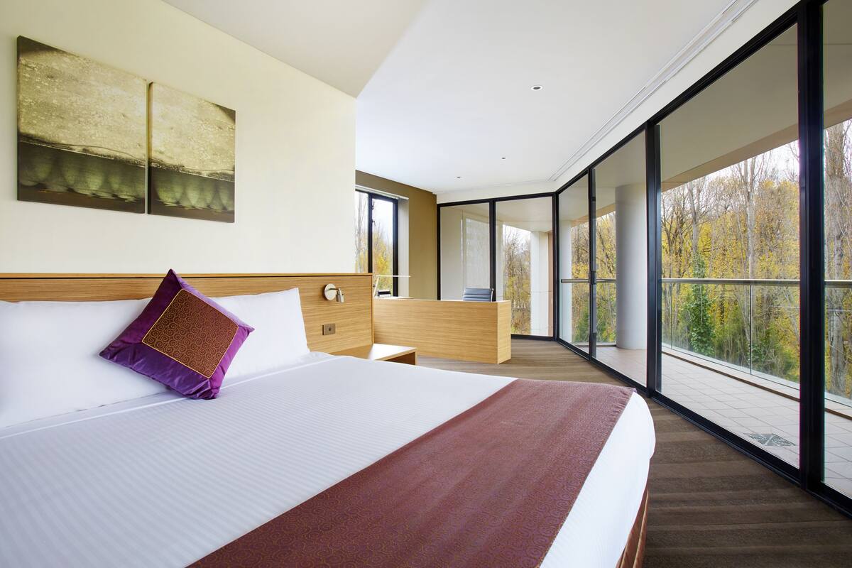 Habitación de hotel moderna con una cama grande, cabecera de madera, un ventanal y un balcón con vistas a árboles.