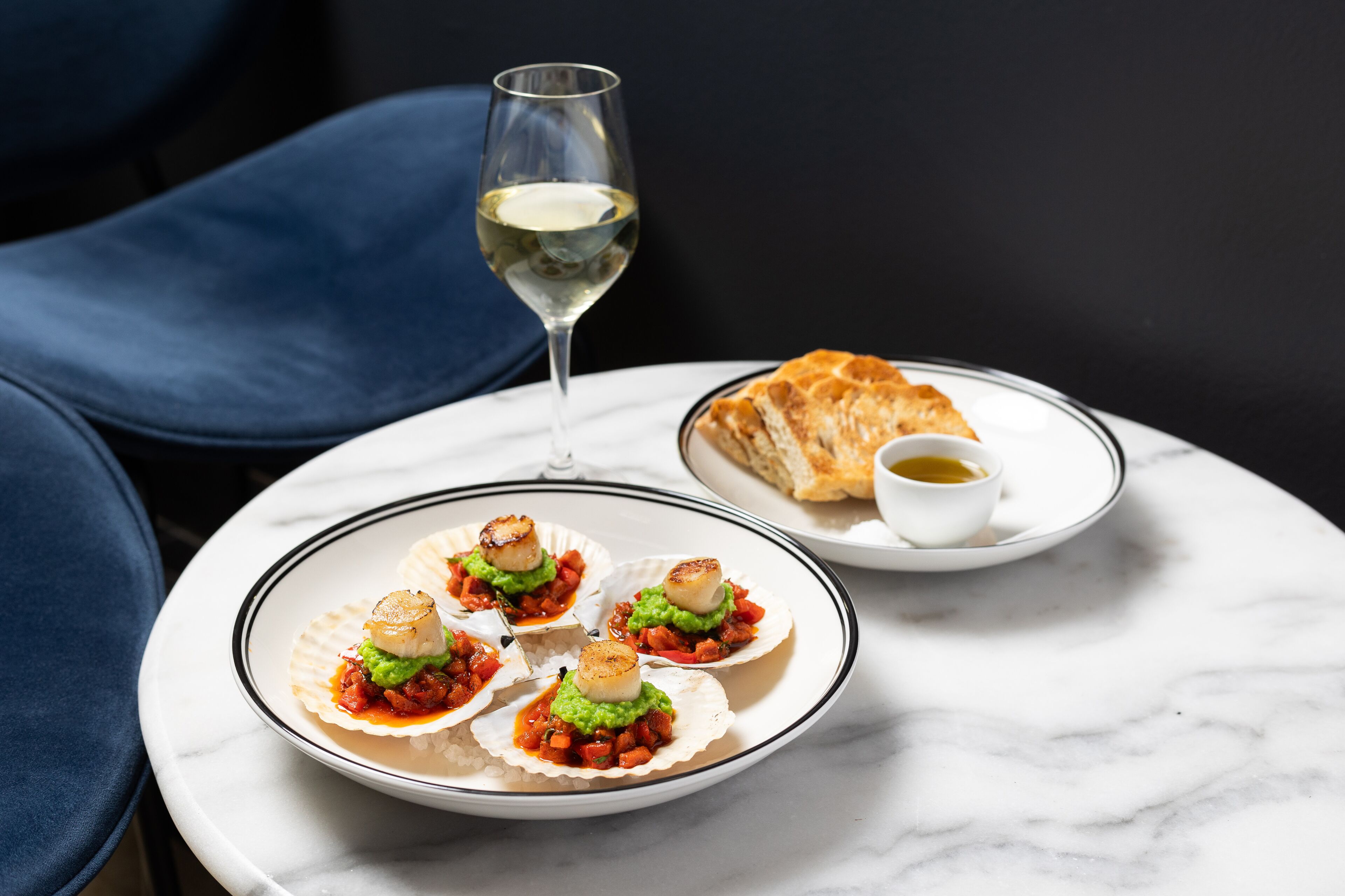 A table with a plate of scallops, a glass of white wine, and a small dish of sauce.