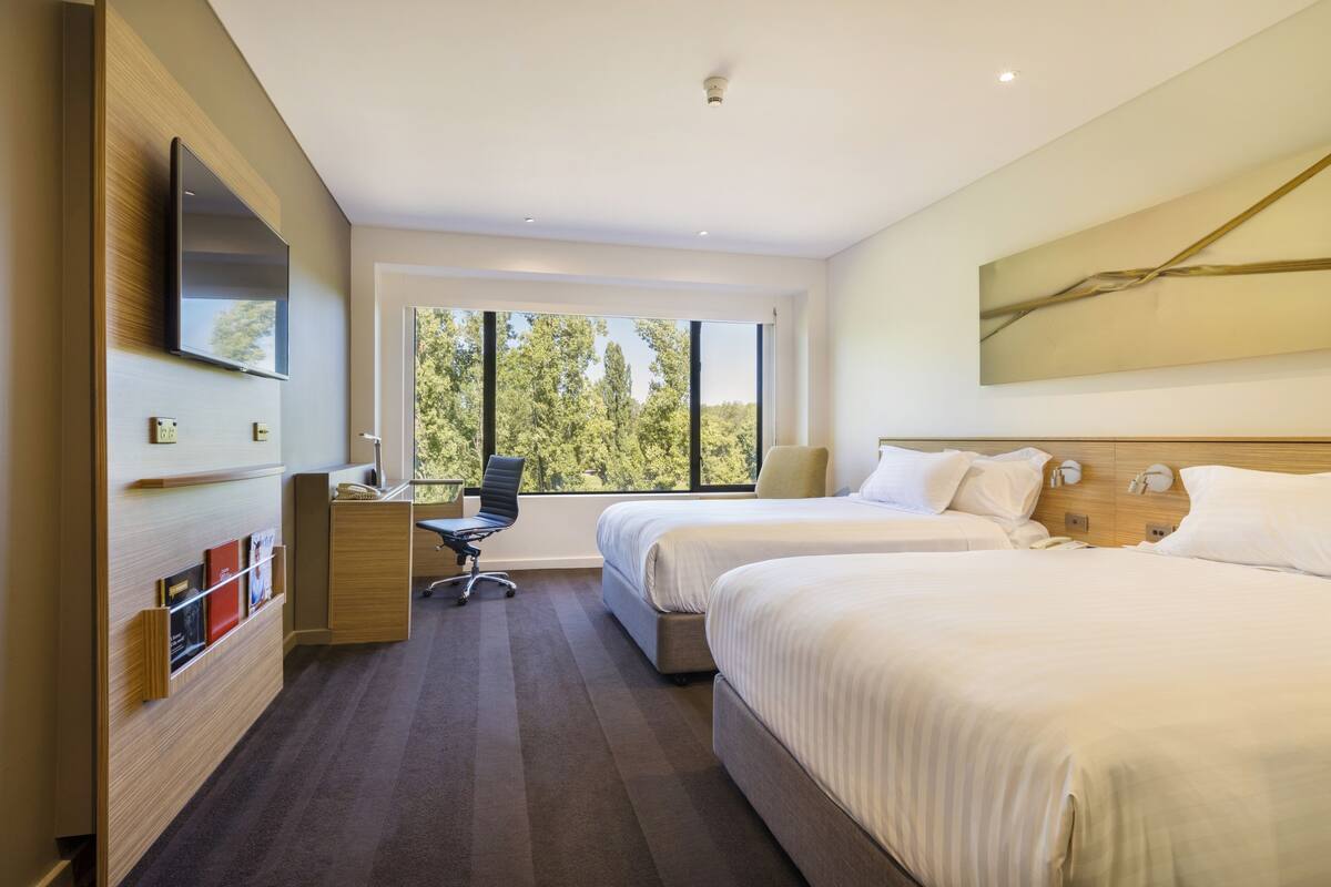 Habitación de hotel con dos camas, un escritorio, una silla, un televisor de pantalla plana y un ventanal con vistas a árboles.