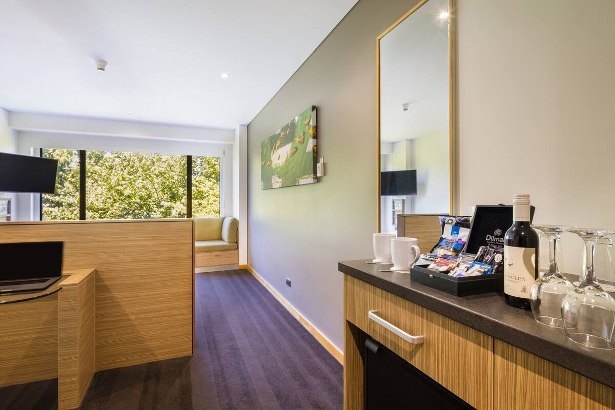 Una habitación de hotel moderna con un escritorio de madera, un televisor de pantalla plana, un espejo grande y un mini bar con copas de vino y una botella.