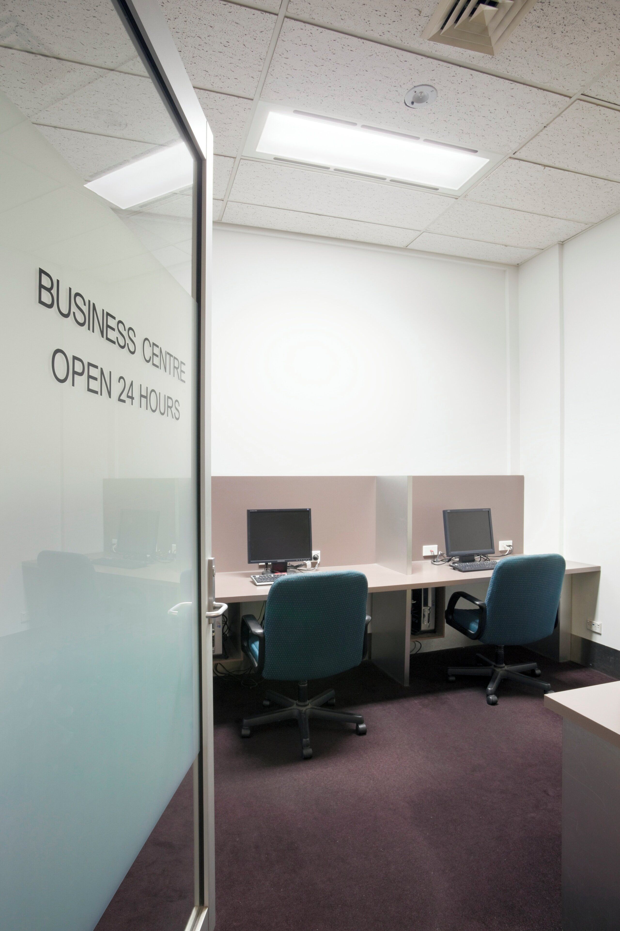Centro de negocios con cubículos, computadoras y una pared de vidrio con el texto 'BUSINESS CENTRE OPEN 24 HOURS'.
