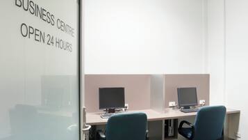 Centro de negocios con cubículos, computadoras y una pared de vidrio con el texto 'BUSINESS CENTRE OPEN 24 HOURS'.