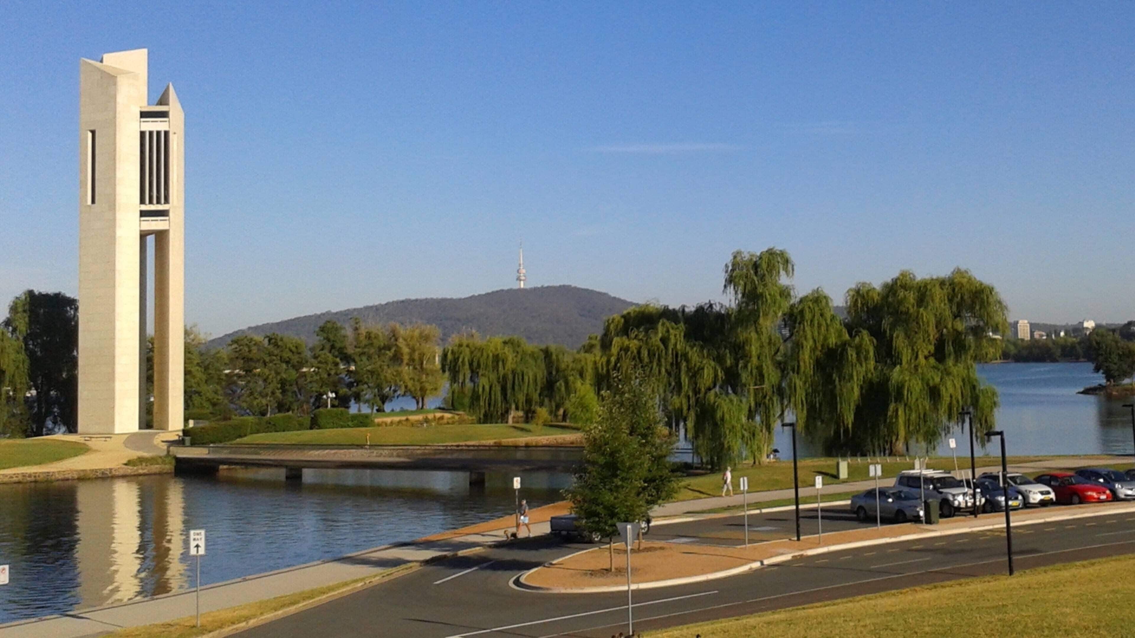 A modern building with a tall, narrow tower, a river, and a hilly landscape in the background.