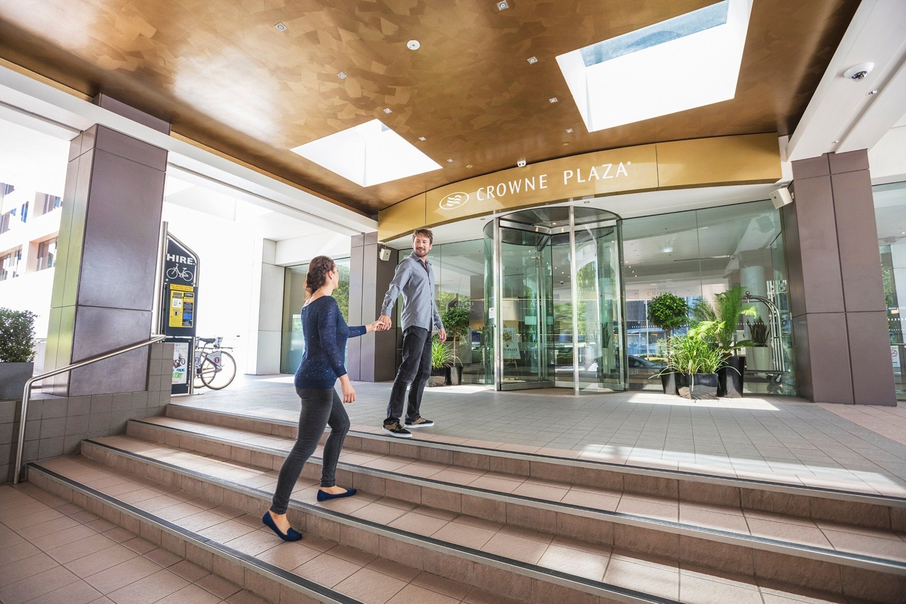 Une entrée moderne avec une porte tournante en verre, une enseigne indiquant « Crowne Plaza » et une affiche pour la location de vélos.