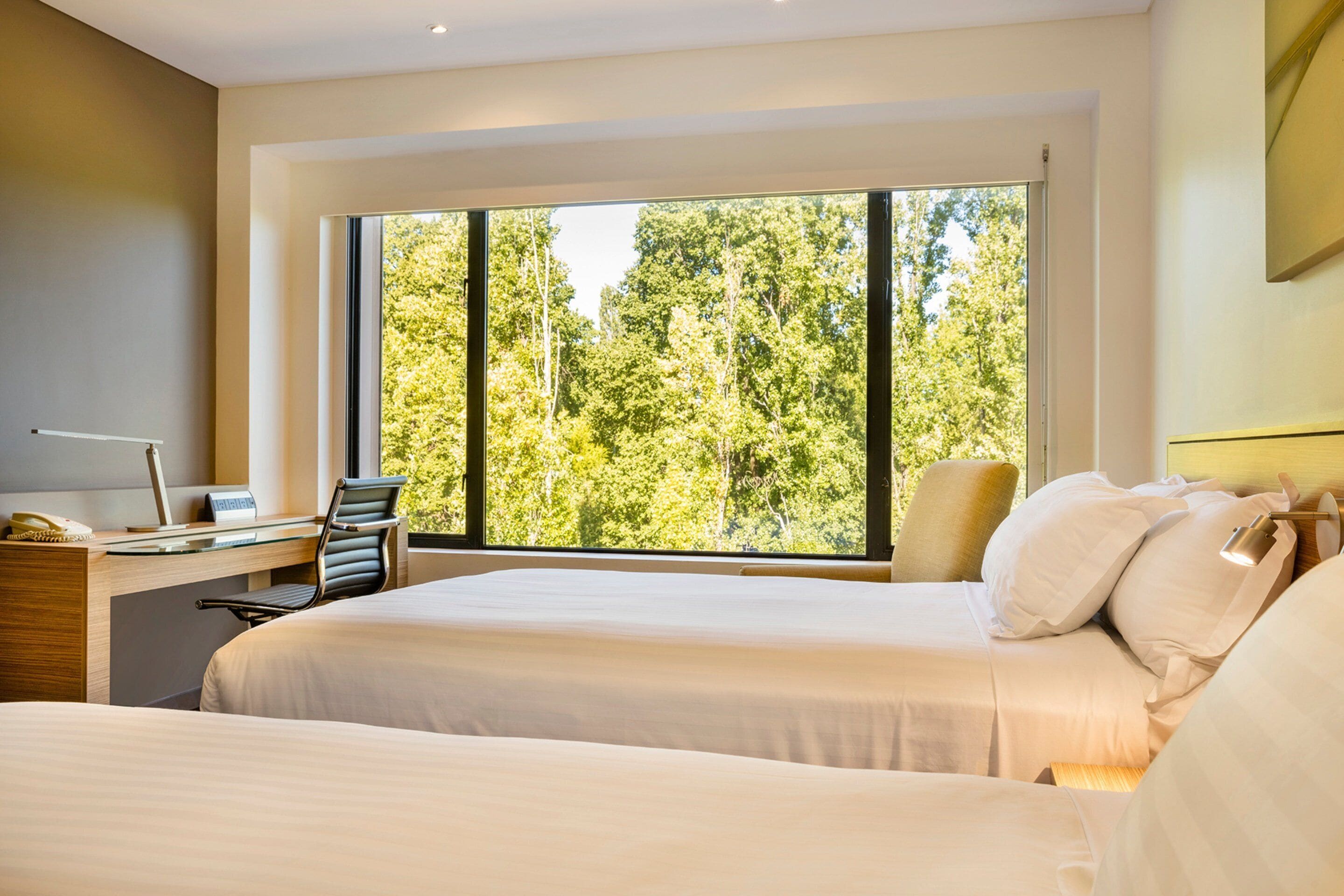 A hotel room with two beds, a desk, and a large window with a view of trees.