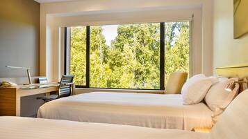 A hotel room with two beds, a desk, and a large window with a view of trees.