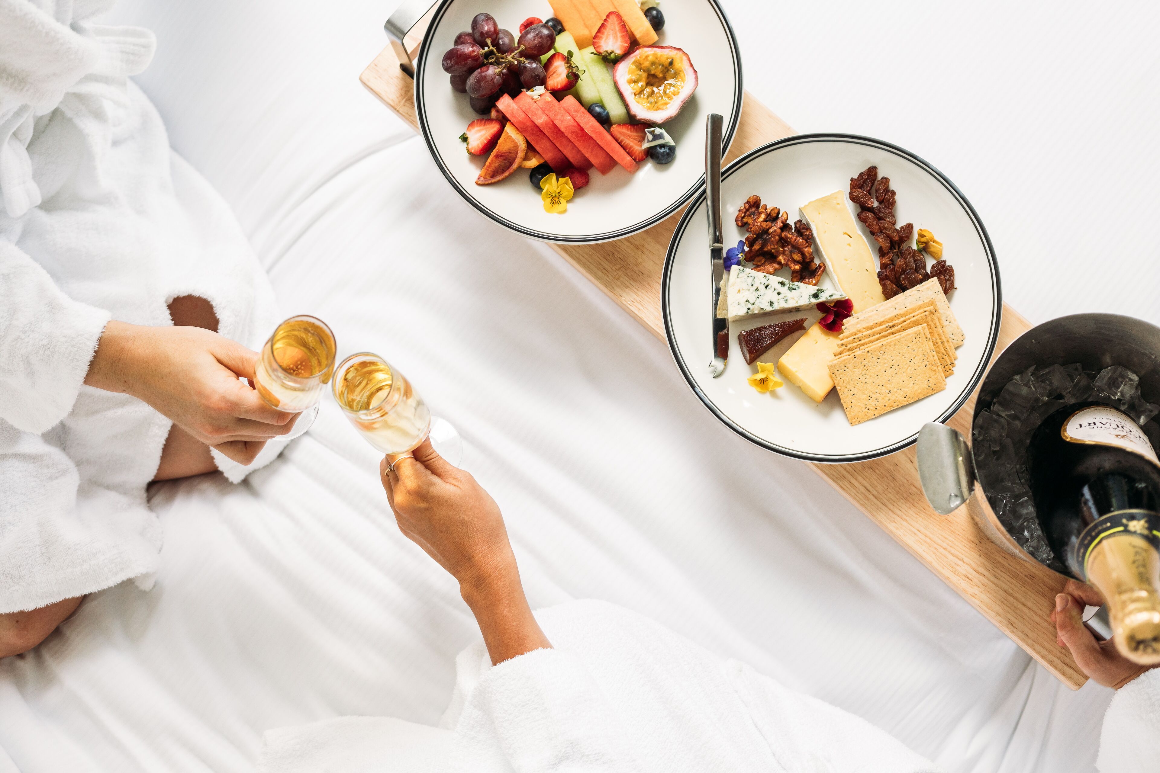 A person in a white bathrobe holding a glass of champagne, with a tray of food and a bottle of champagne on a wooden table.