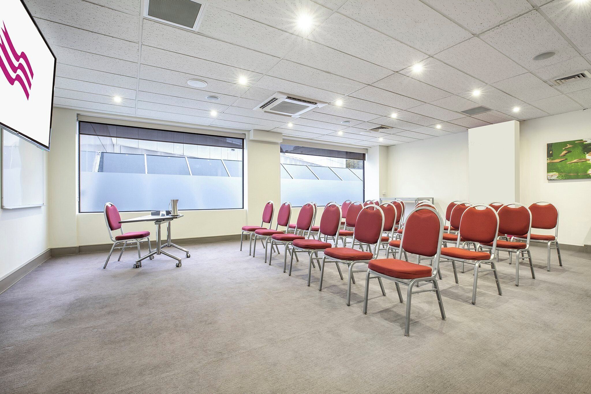 Un salón de conferencias con filas de sillas rojas, una pantalla de proyector y ventanas amplias.
