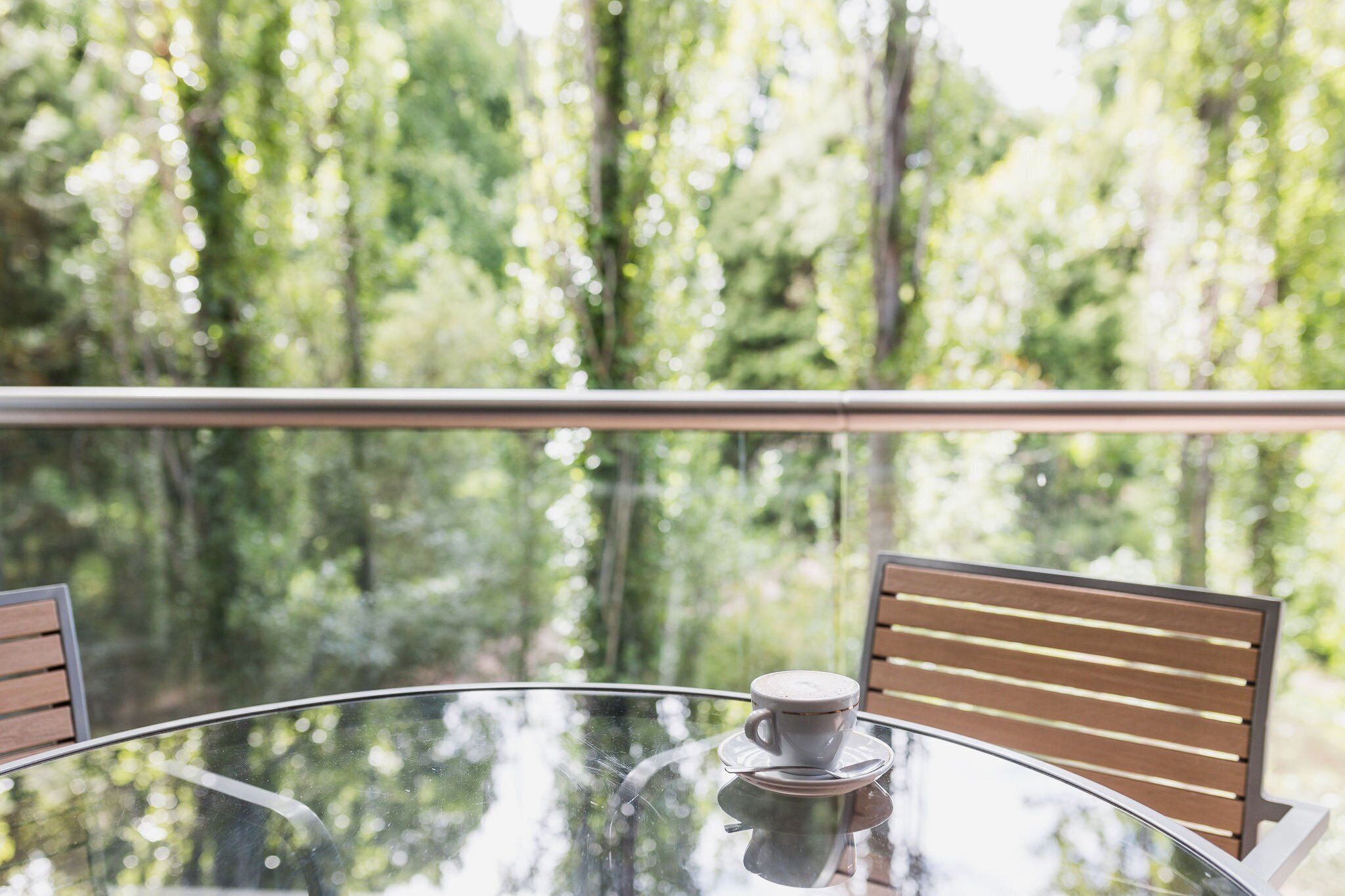 A round glass table with a cup of coffee, two wooden chairs, and a view of a forest through a balcony railing.