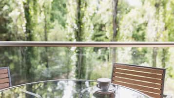 A round glass table with a cup of coffee, two wooden chairs, and a view of a forest through a balcony railing.