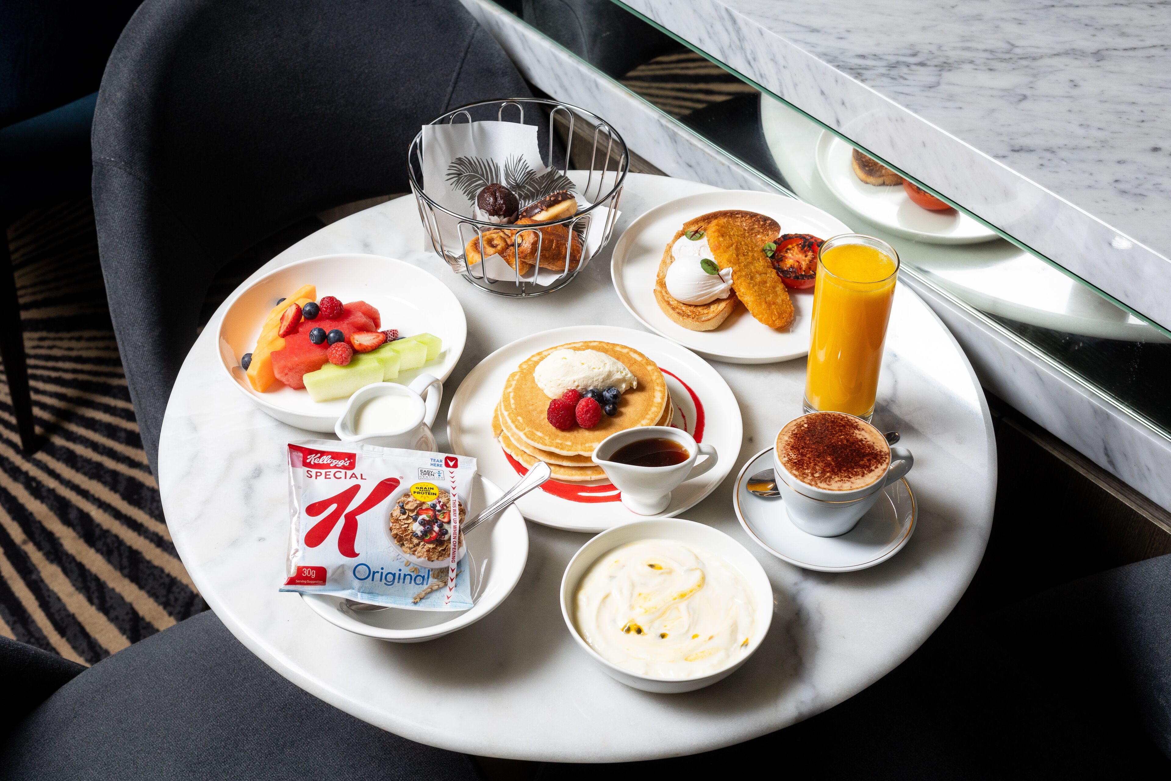 A breakfast tray with pancakes, fruit, yogurt, and a juice glass on a marble table.