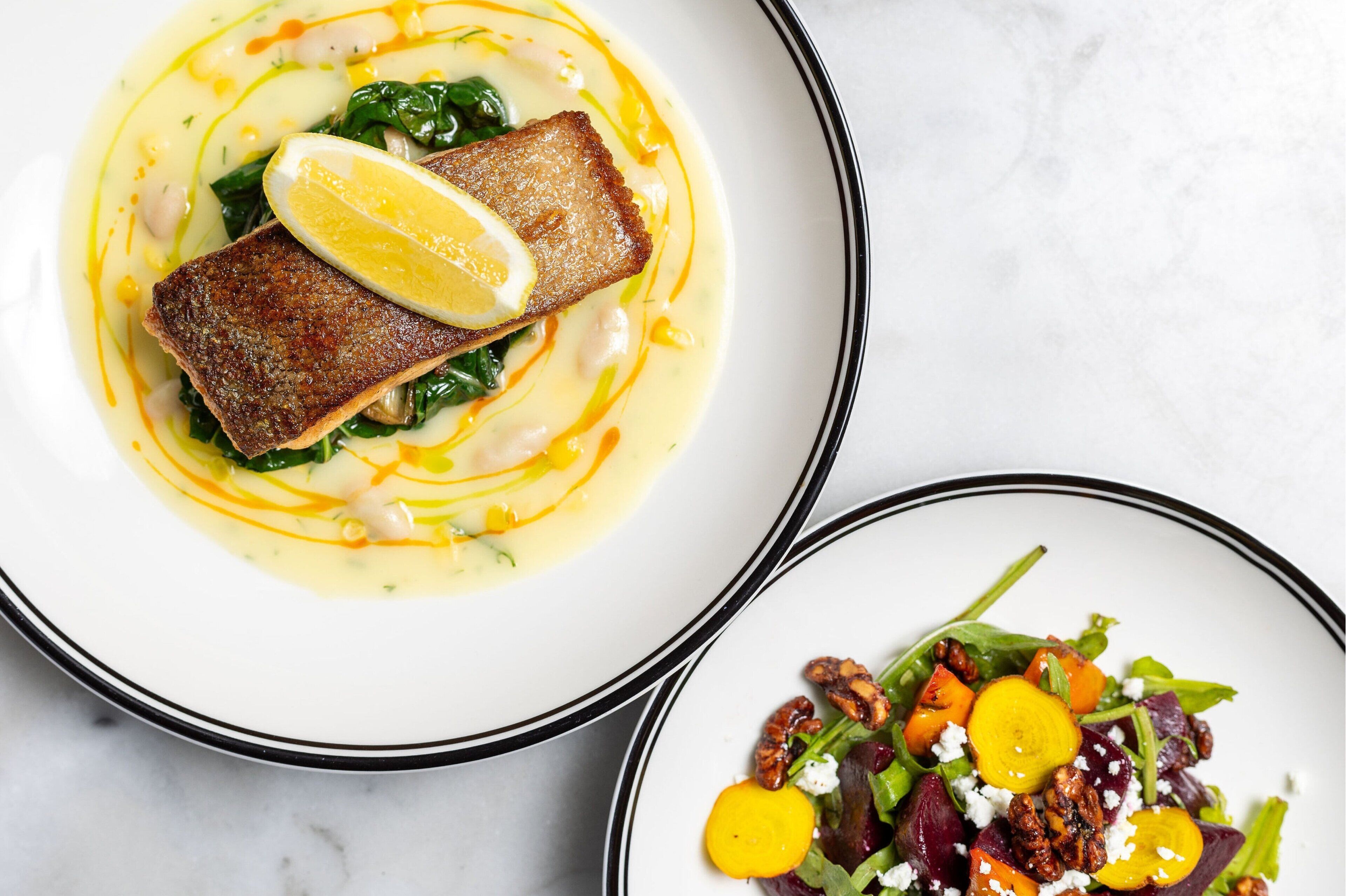 A plate with a piece of fish and greens, and another plate with a salad.
