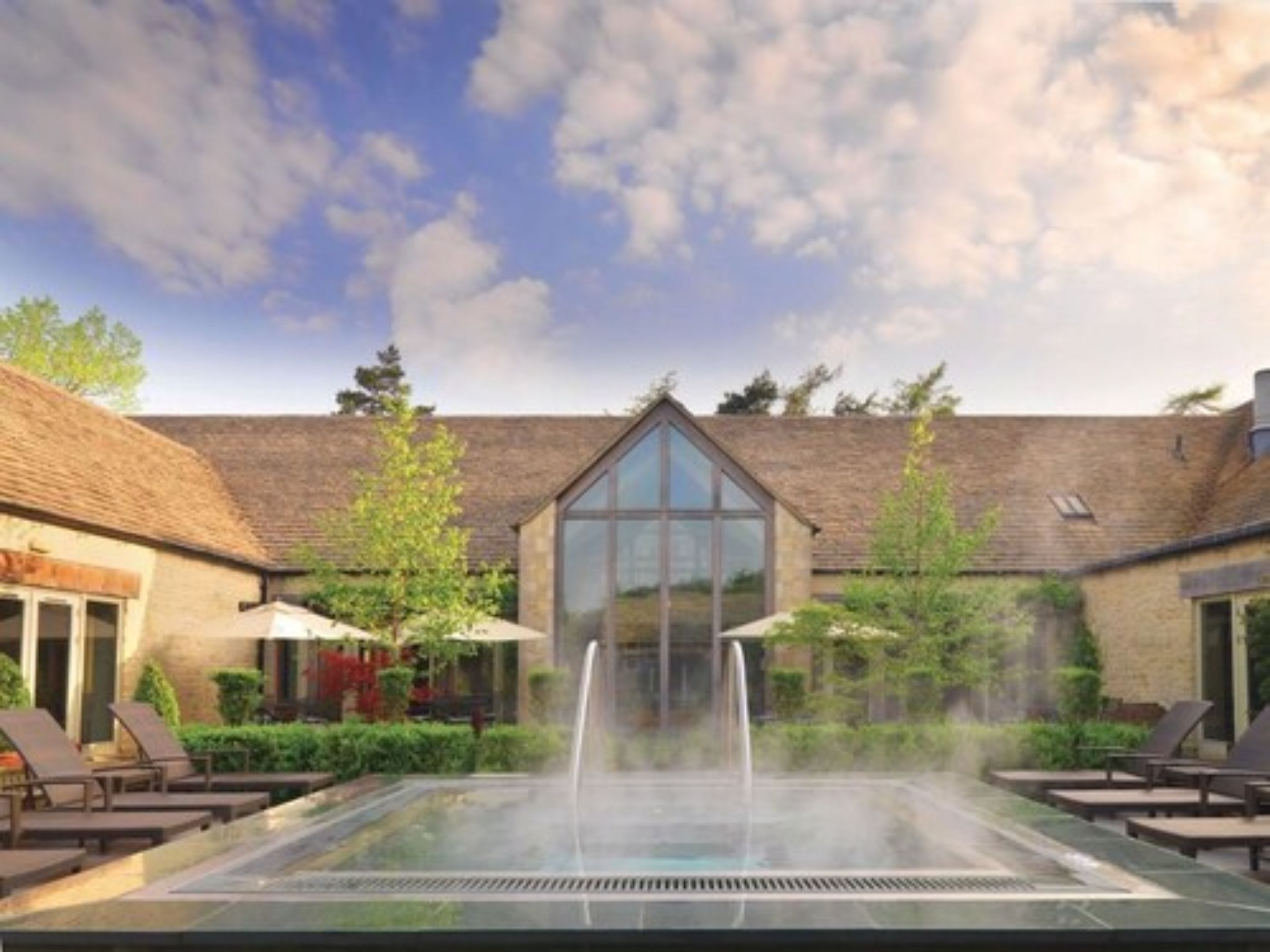 A spa area with a glass-enclosed hot tub, lounge chairs, and a building with a sloped roof in the background.