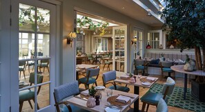 A spacious dining area with multiple tables and chairs, large windows, and indoor plants.