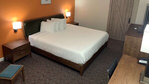 Une chambre d’hôtel avec un grand lit, deux lampes de chevet, une tête de lit en bois, une table de chevet avec des tiroirs, une petite table avec un coussin, un bureau avec un ordinateur et un téléphone, et une porte coulissante.