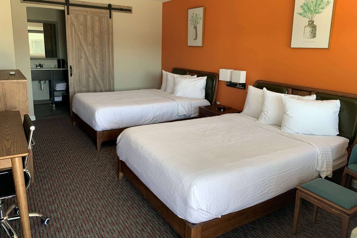 Hotelkamer met twee bedden, een bureau, een stoel, een klein tafeltje, een nachtkastje en een zichtbare badkamer door een geopende deur.