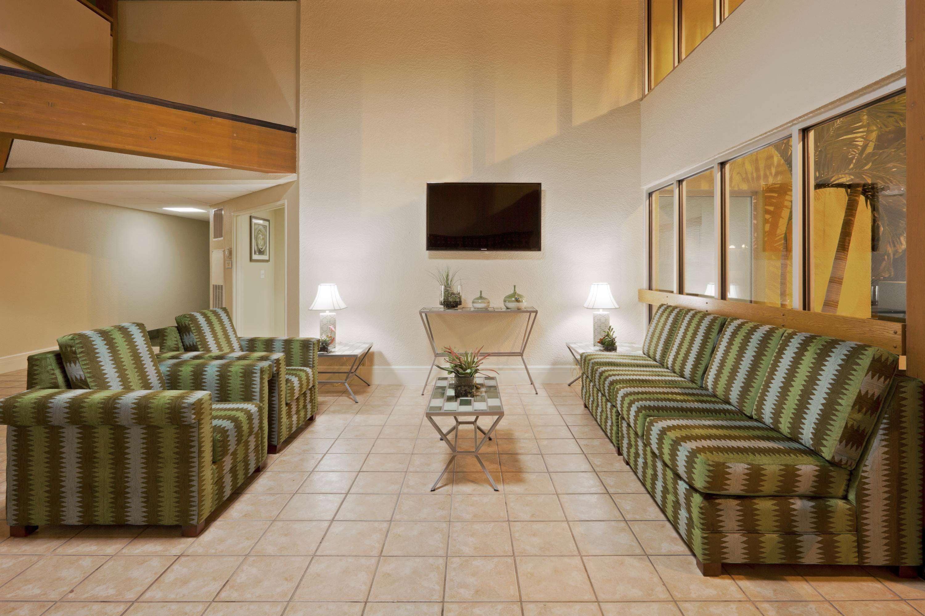 A spacious living room with a high ceiling, a flat-screen TV mounted on the wall, and a large window with a view of palm trees.