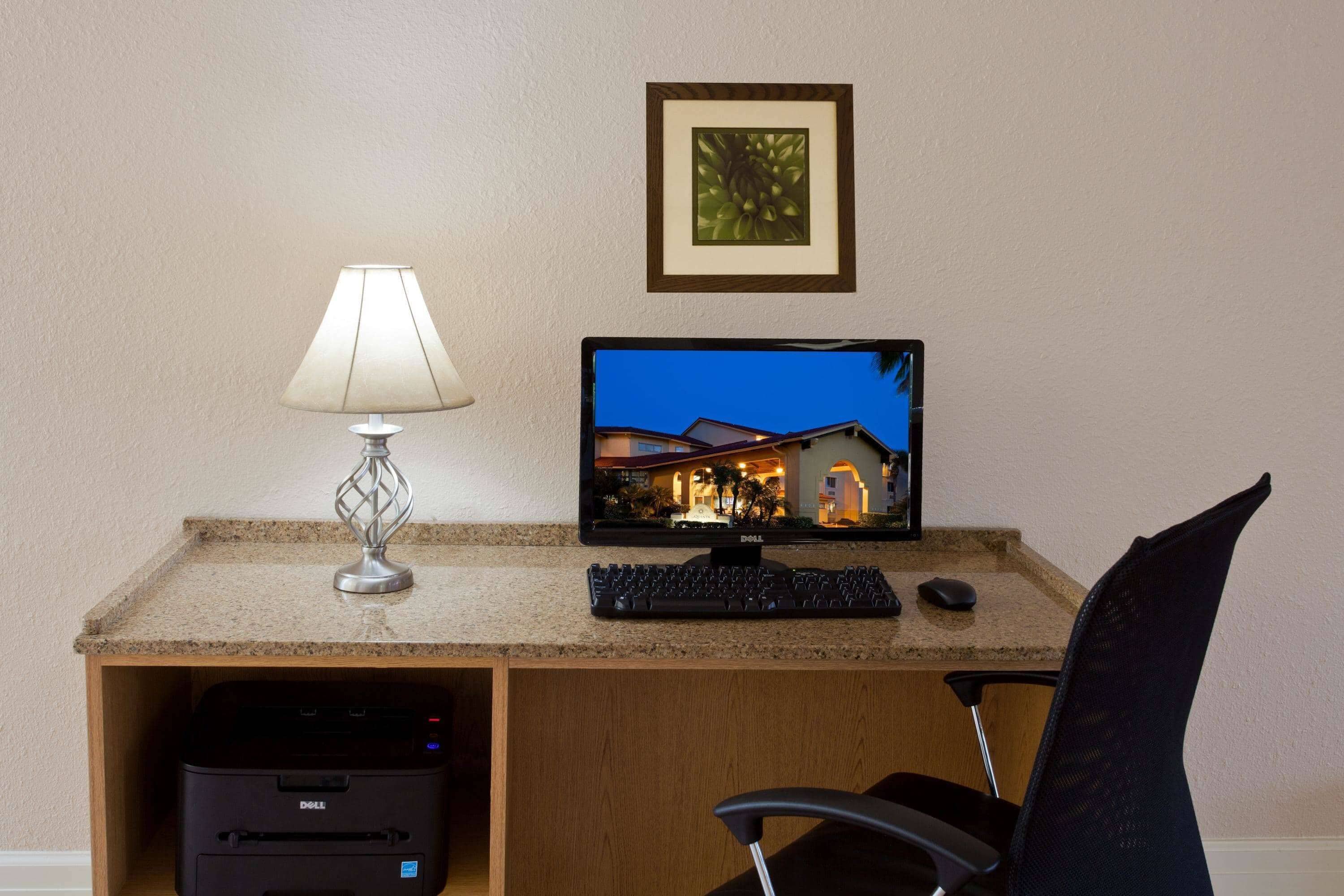 A hotel room desk with a computer, keyboard, mouse, and lamp.