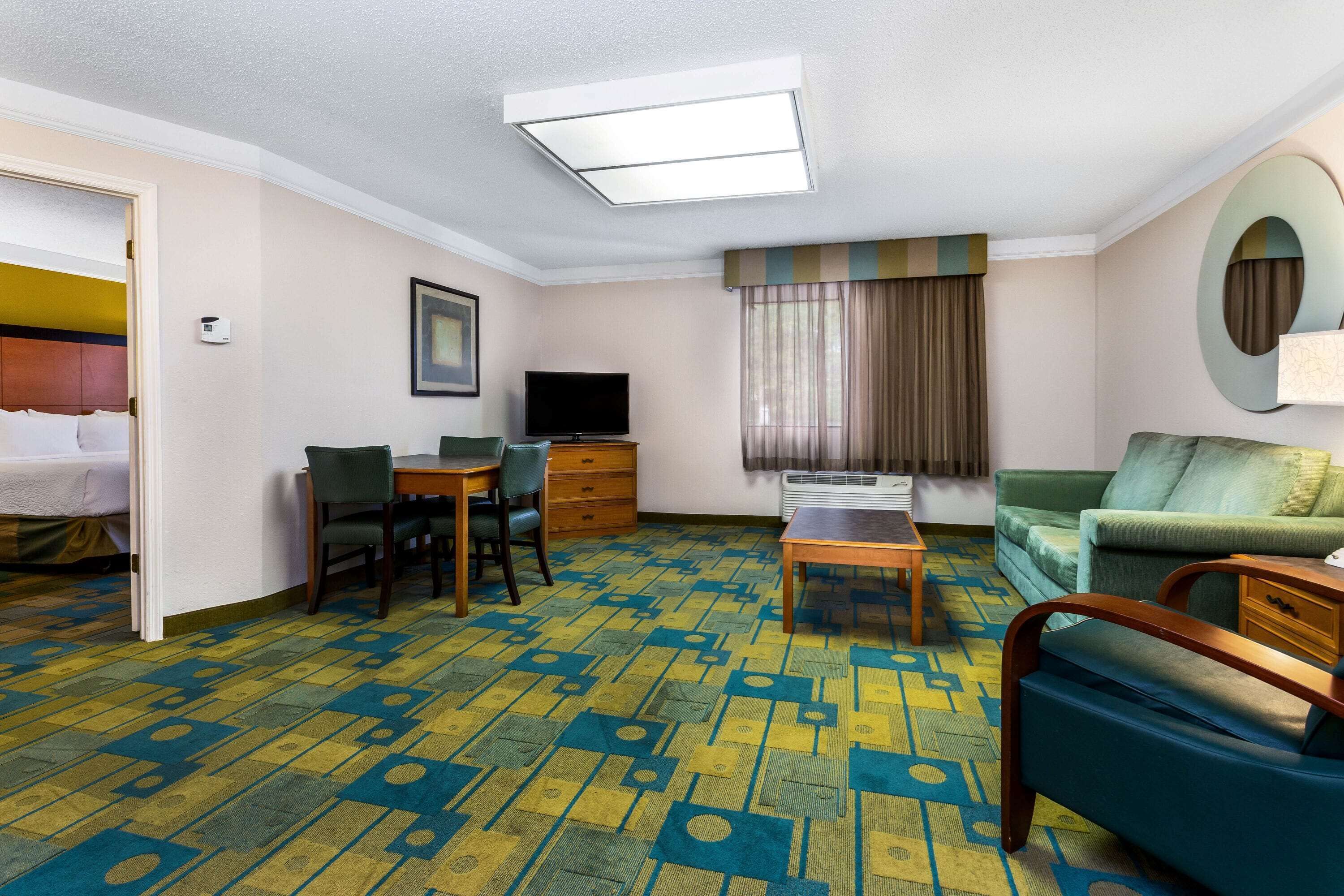 A hotel room with a bed, a desk with a chair, a sofa, a television, and a patterned carpet.