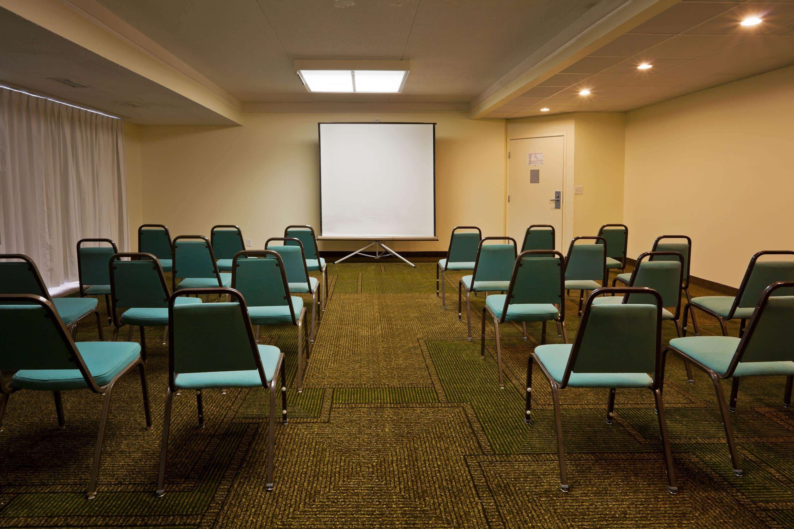 Ein Konferenzraum mit Reihen grüner Stühle, einem Teppichboden, einer weißen Projektionsleinwand und einer Tür im Hintergrund.