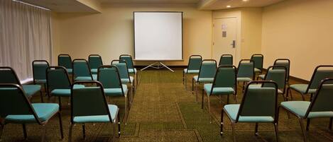A conference room with rows of green chairs, a carpeted floor, a white projection screen, and a door in the background.