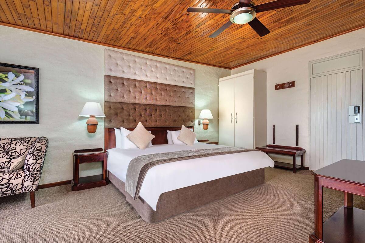 Un dormitorio con techo de madera, cabecera capitonada, cama grande, dos lámparas de noche, una mesita de noche, una mesa de madera y un cuadro enmarcado en la pared.