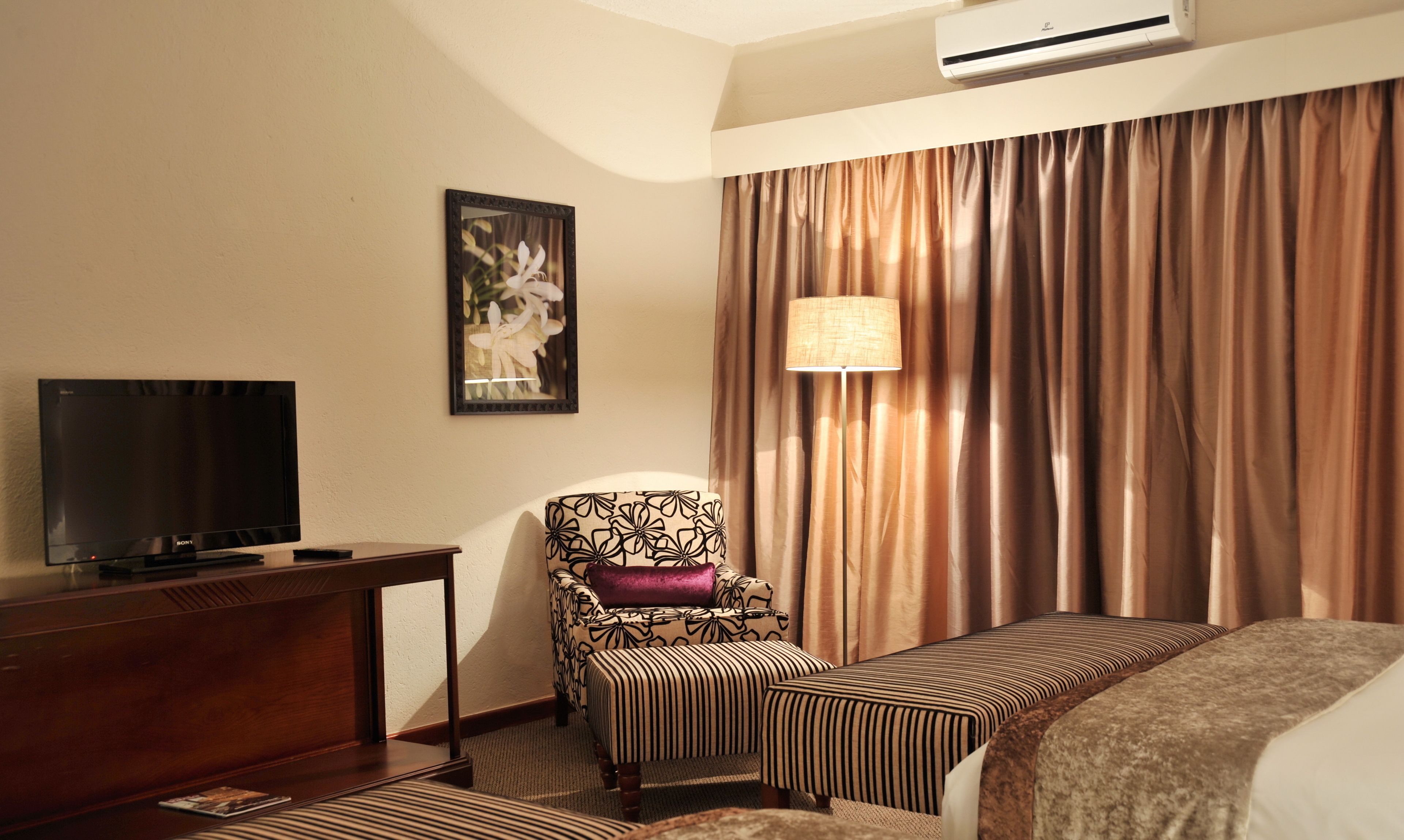 A hotel room with a bed, a chair, a desk with a television, a lamp, and a large window with curtains.