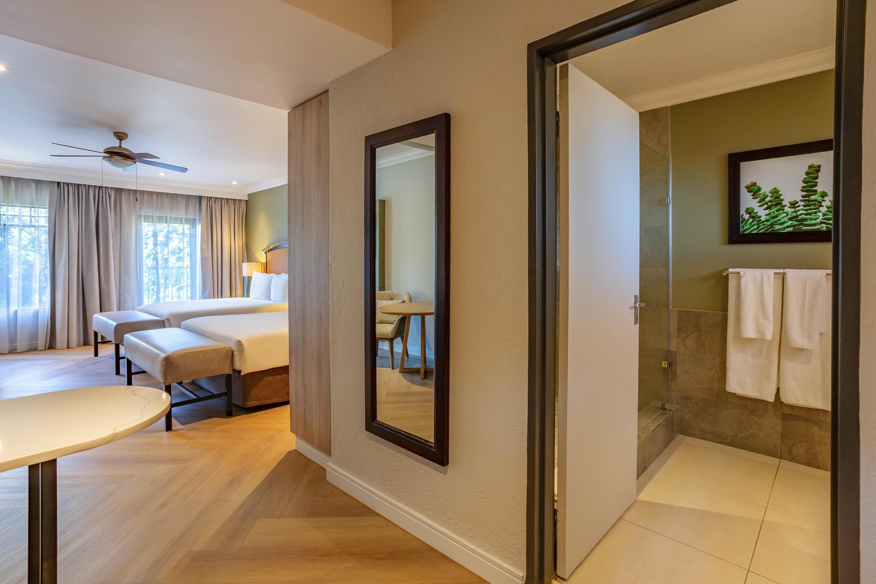 A hotel room with a bed, a desk, a chair, a mirror, and a bathroom with a shower and a towel rack.