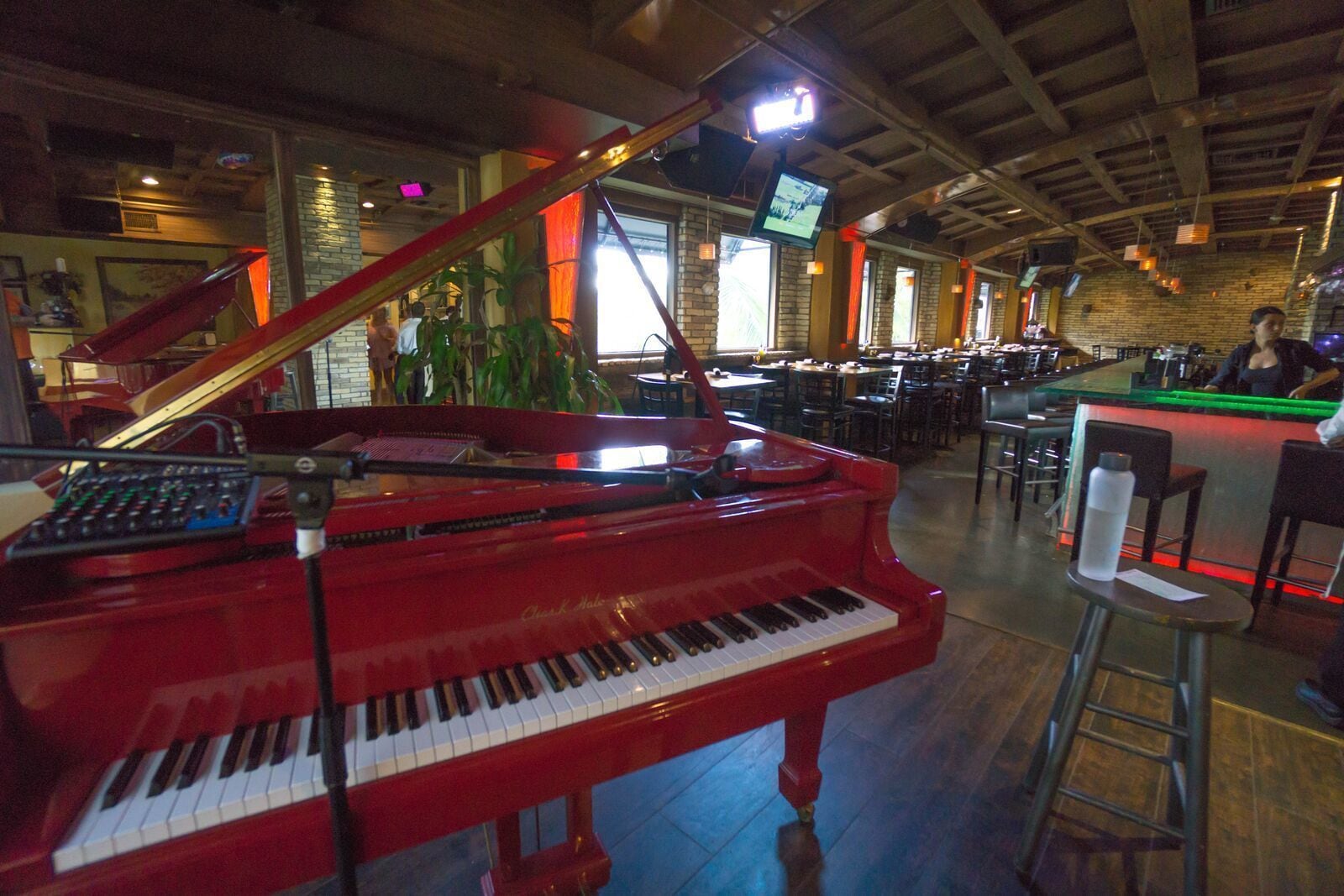 A red grand piano in a spacious indoor area with tables and chairs, a bar counter, and a television mounted on the wall.