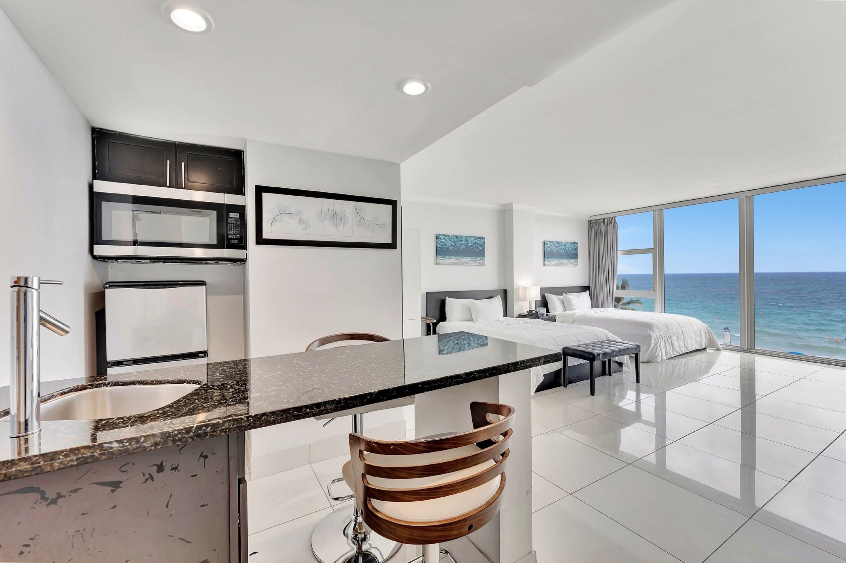 A modern hotel room with a kitchen area, a bed with a view of the ocean, and a bar with stools.