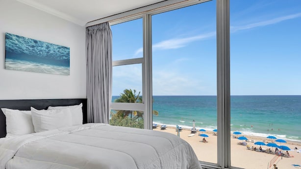 Une chambre avec une grande fenêtre donnant sur une plage, un lit avec une literie blanche et un tableau représentant l’océan accroché au mur.