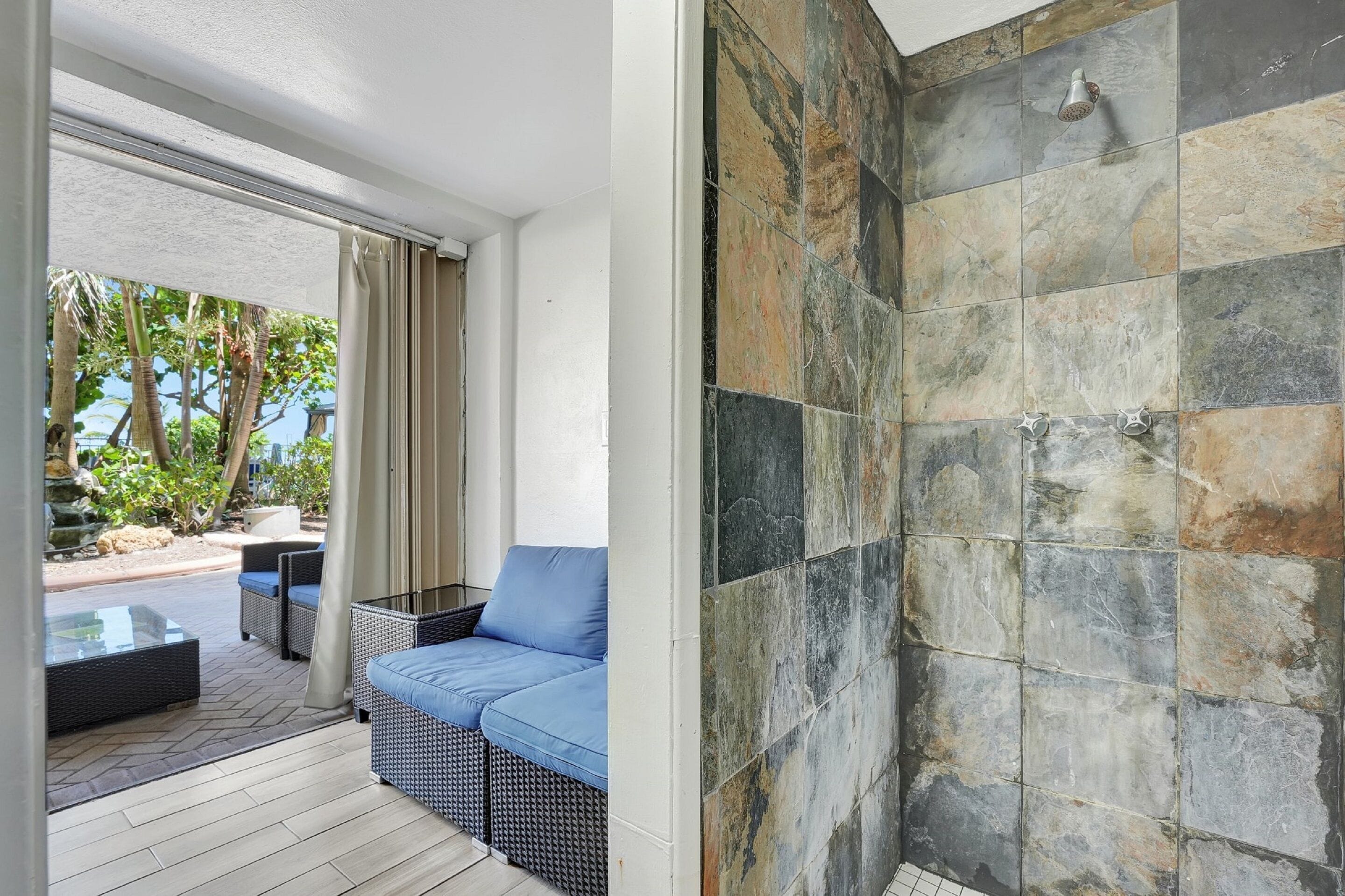 A room with a sofa and a glass table, a tiled shower wall, and a view of trees and a rock formation outside.