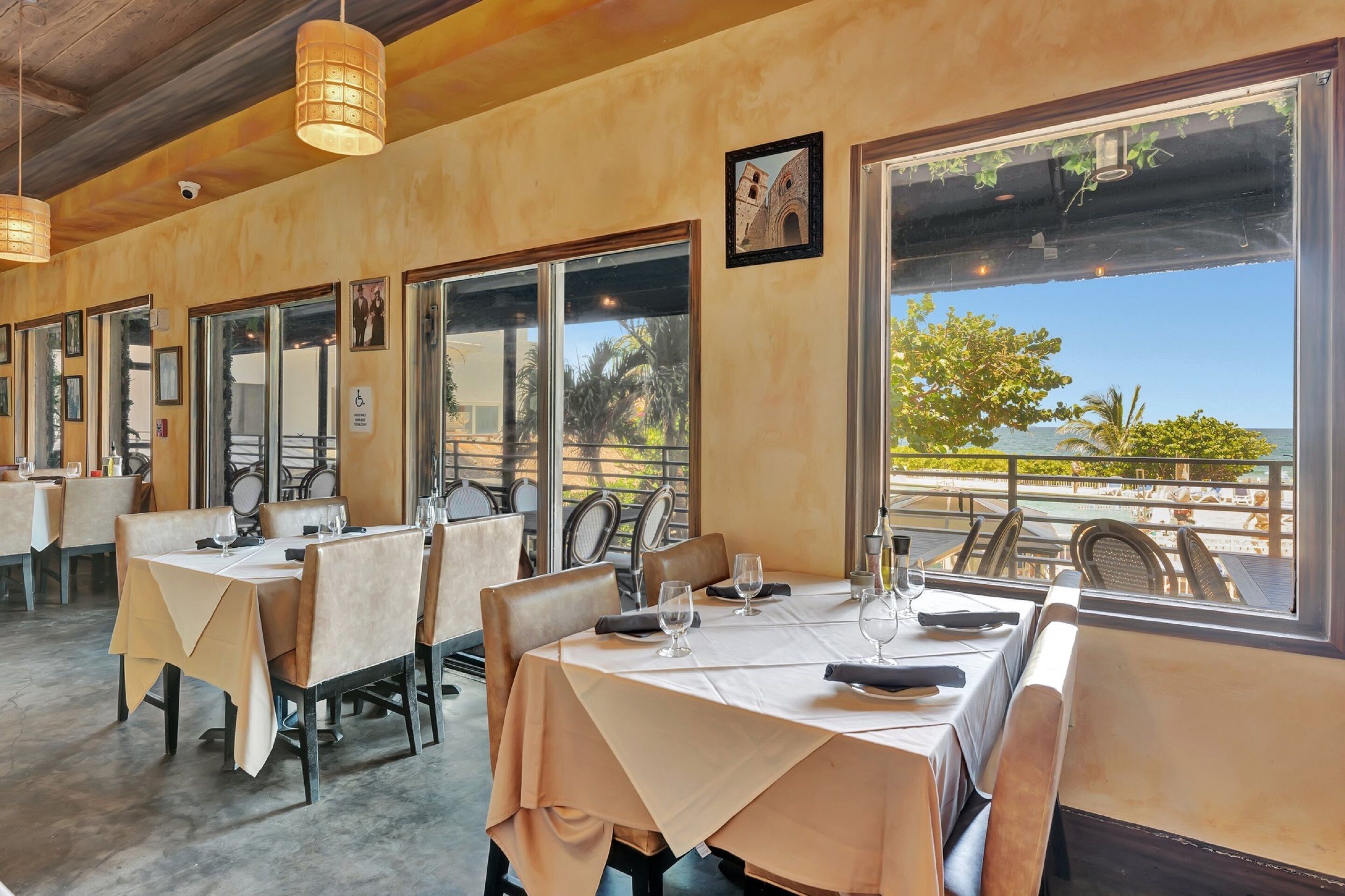 A restaurant with tables set for dining, large windows with a view of the sea, and a wooden ceiling.