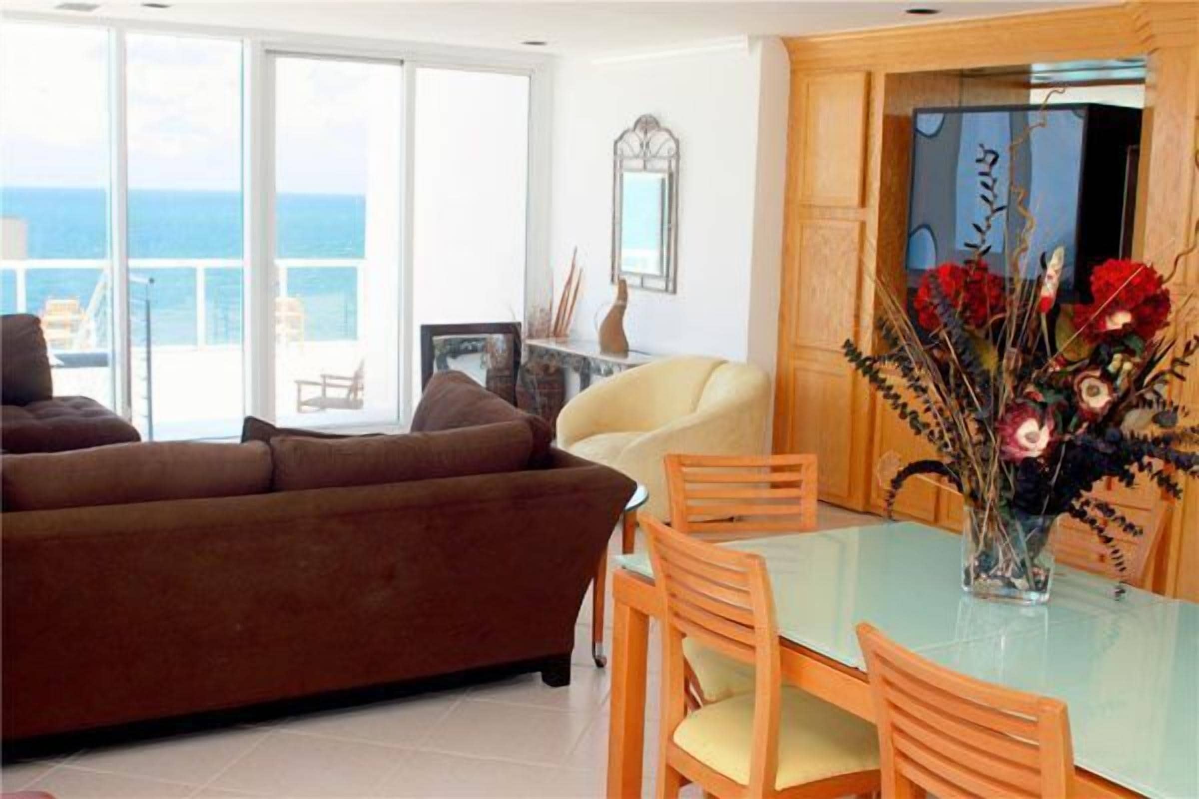 Ein Wohnzimmer mit einem braunen Sofa, einem Glastisch mit Holzstühlen und einem Blumenstrauß.