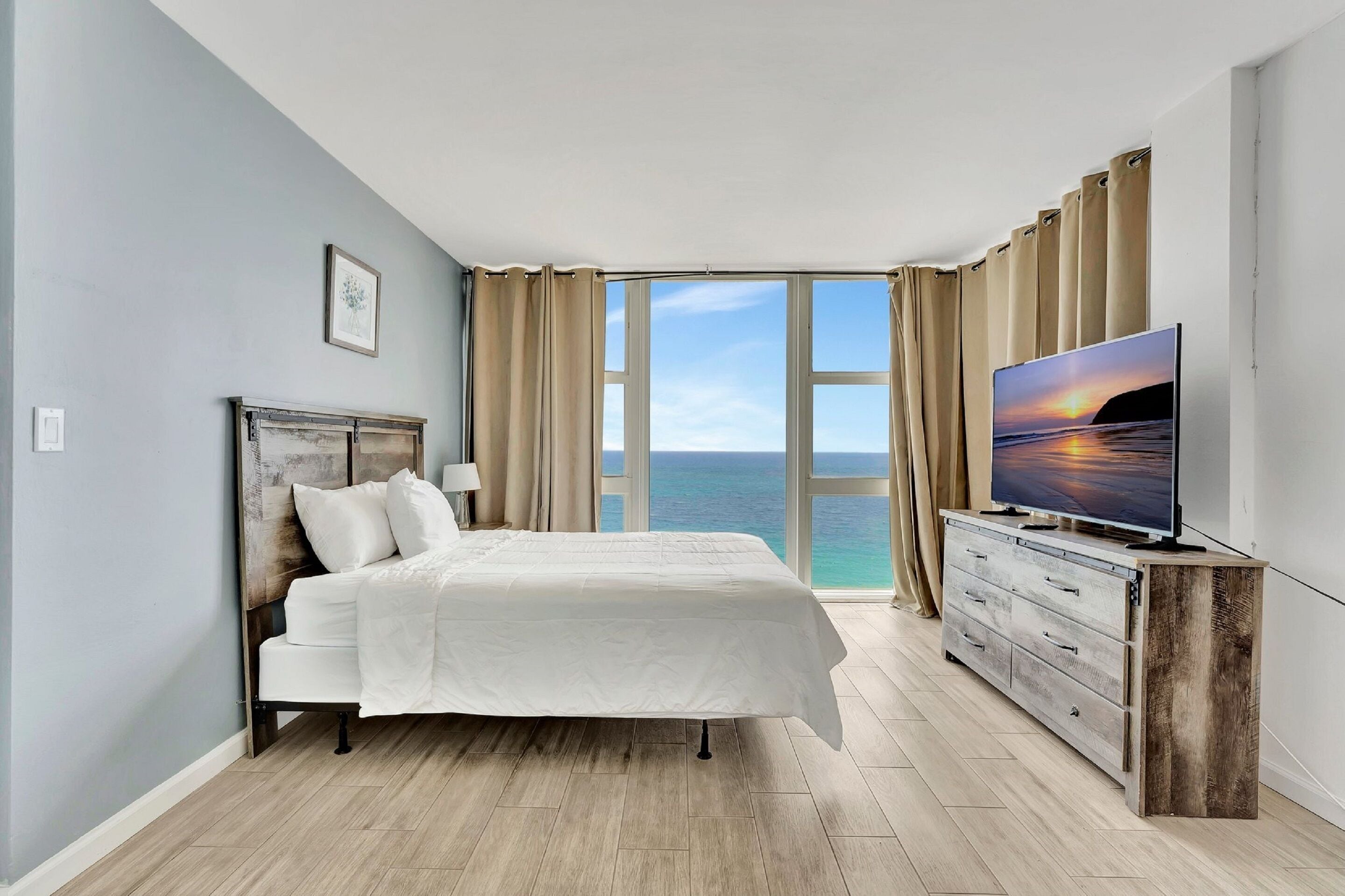 Un dormitorio con cama, televisión, cómoda y una ventana grande con vista al océano.