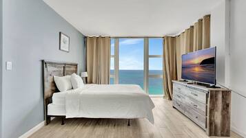 Un dormitorio con cama, televisión, cómoda y una ventana grande con vista al océano.