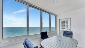 Une pièce avec une table ronde et deux chaises, de grandes fenêtres donnant sur la mer et une porte menant à un balcon.