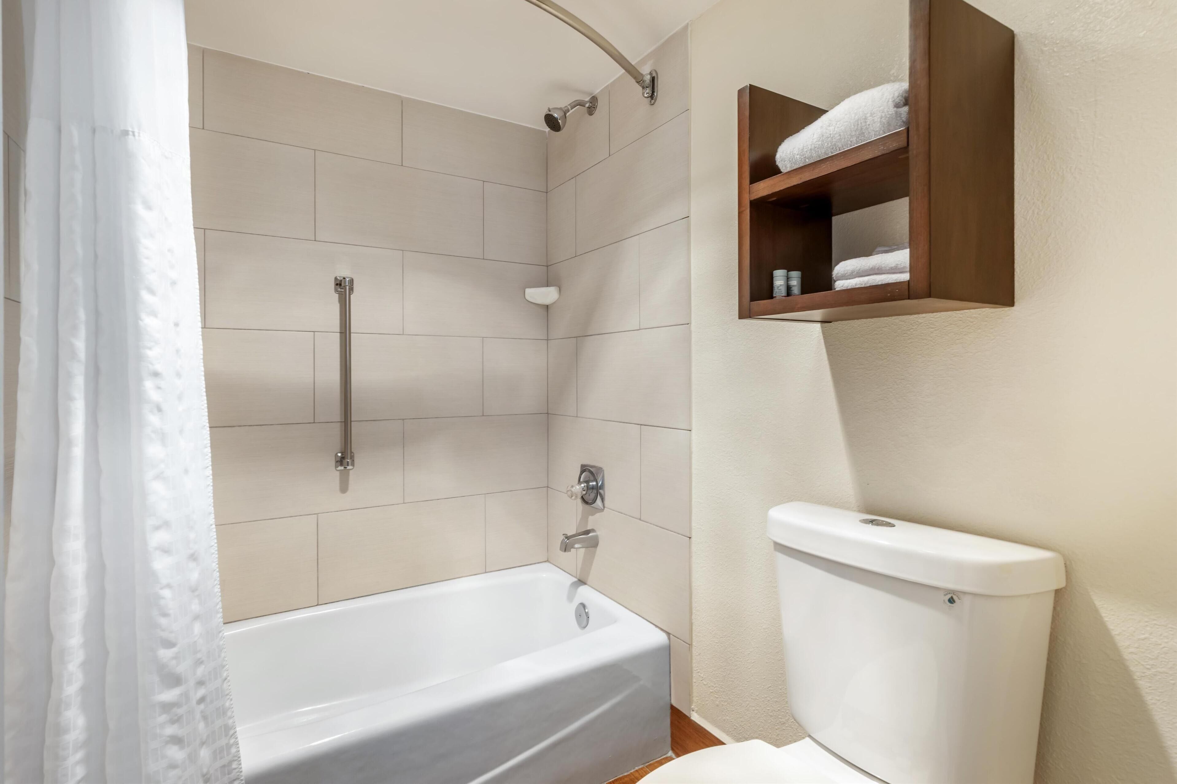 A bathroom with a bathtub, a shower, and a toilet. There is a wooden shelf with towels above the toilet.