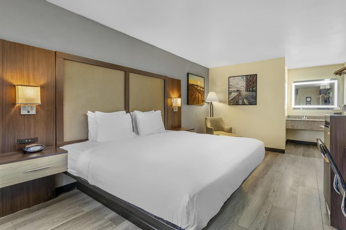 A hotel room with a large bed, a nightstand with a lamp, a chair, a small table, a mirror, and a painting on the wall.