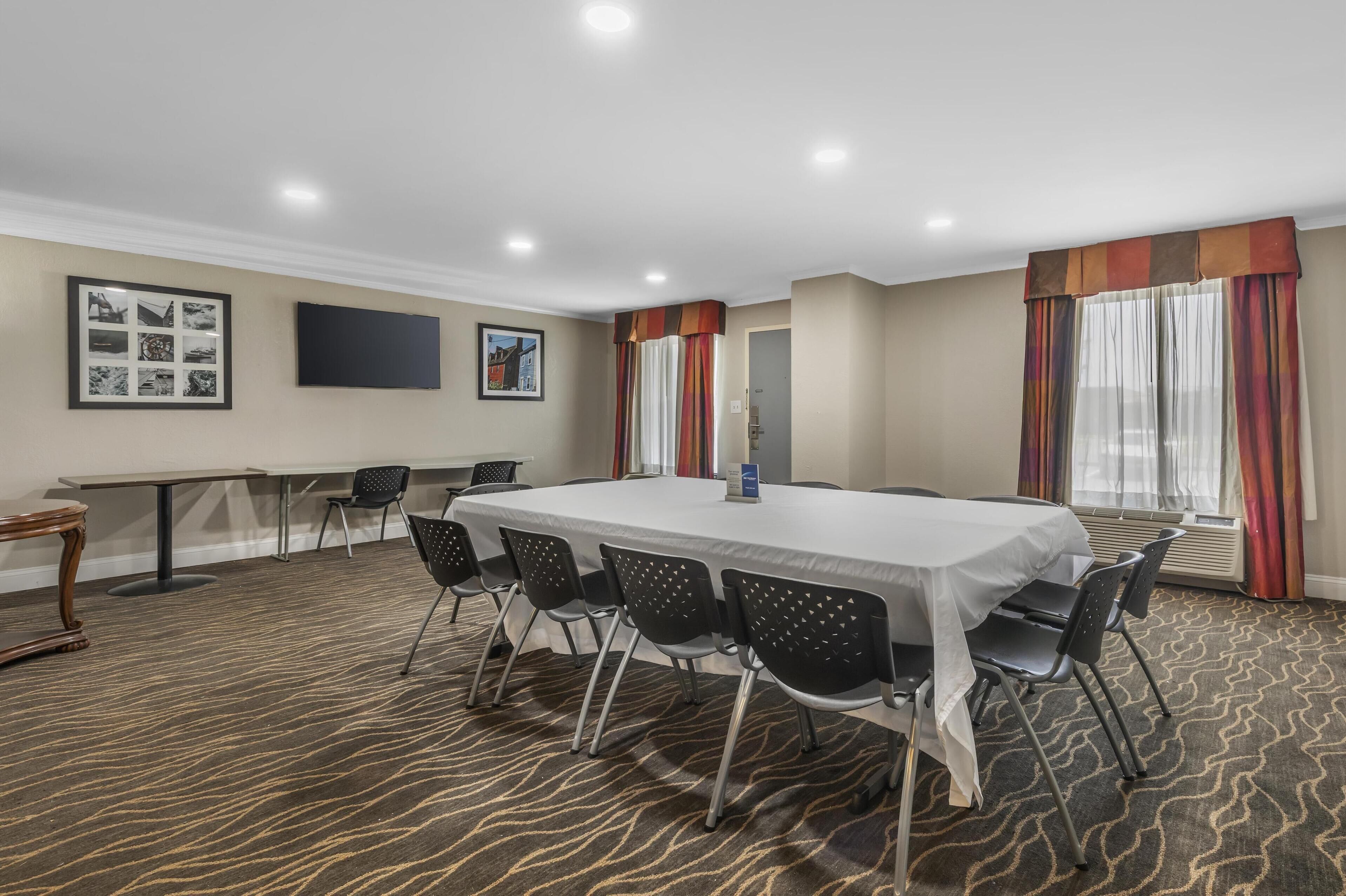 A conference room with a long table, black chairs, framed pictures on the wall, and large windows with curtains.