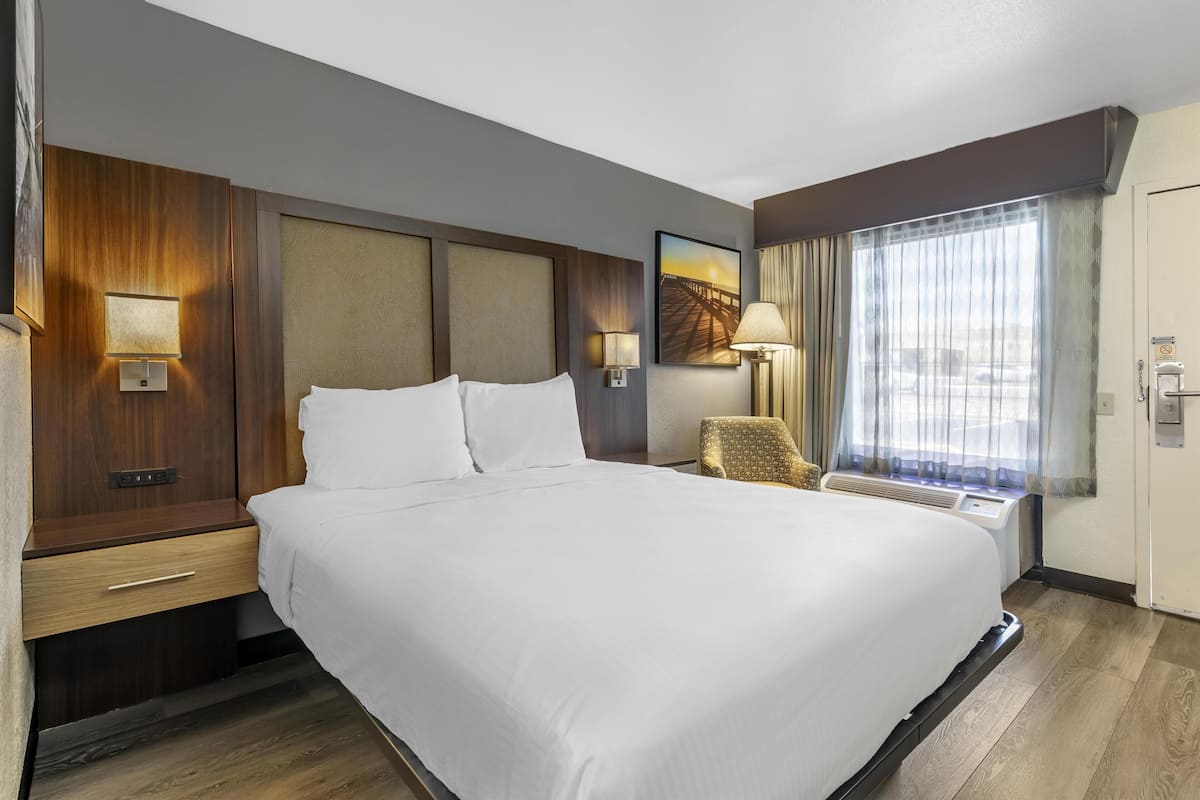 A hotel room with a large bed, wooden headboard, bedside tables, a chair, a painting, and a window with curtains.