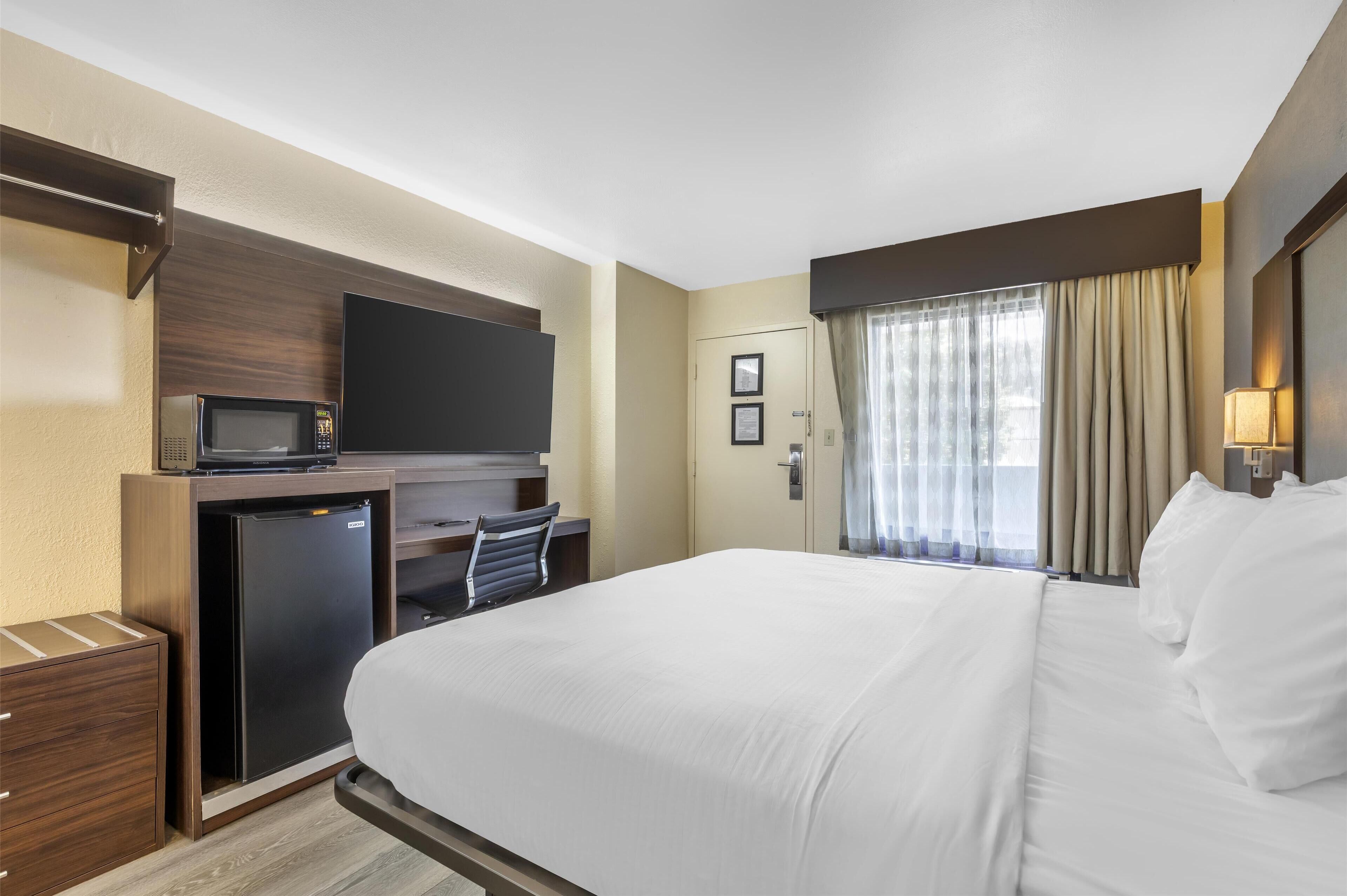 A hotel room with a bed, a desk with a television, a refrigerator, and a window with curtains.