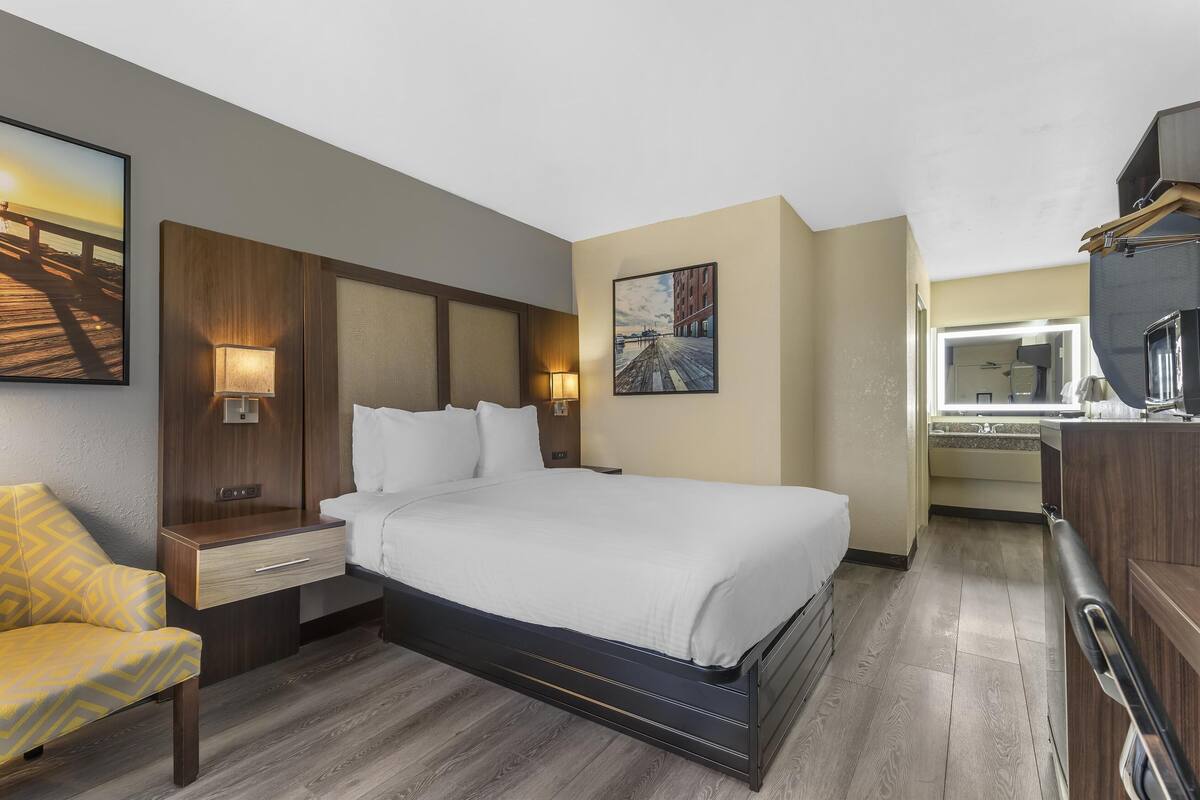 A hotel room with a bed, bedside table, lamp, chair, and artwork on the walls.
