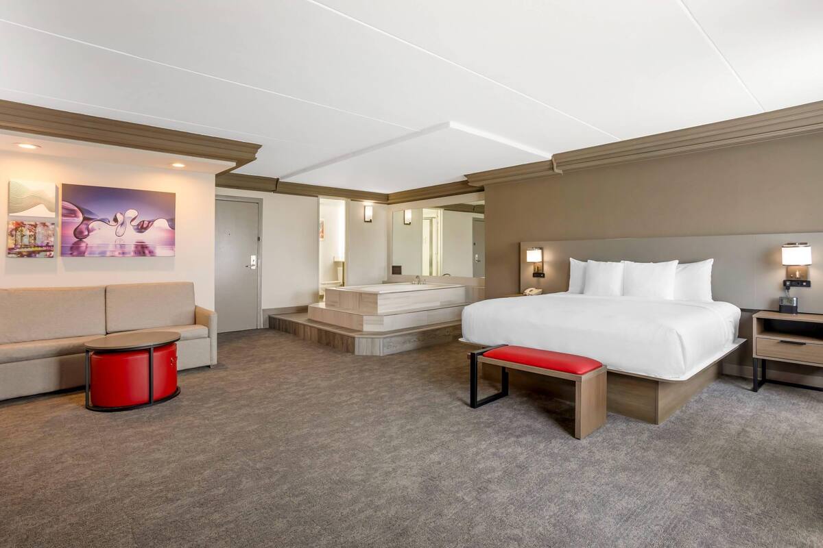 Une chambre d’hôtel avec un lit, un canapé, une petite table rouge, un banc, un miroir et une salle de bain avec une baignoire.